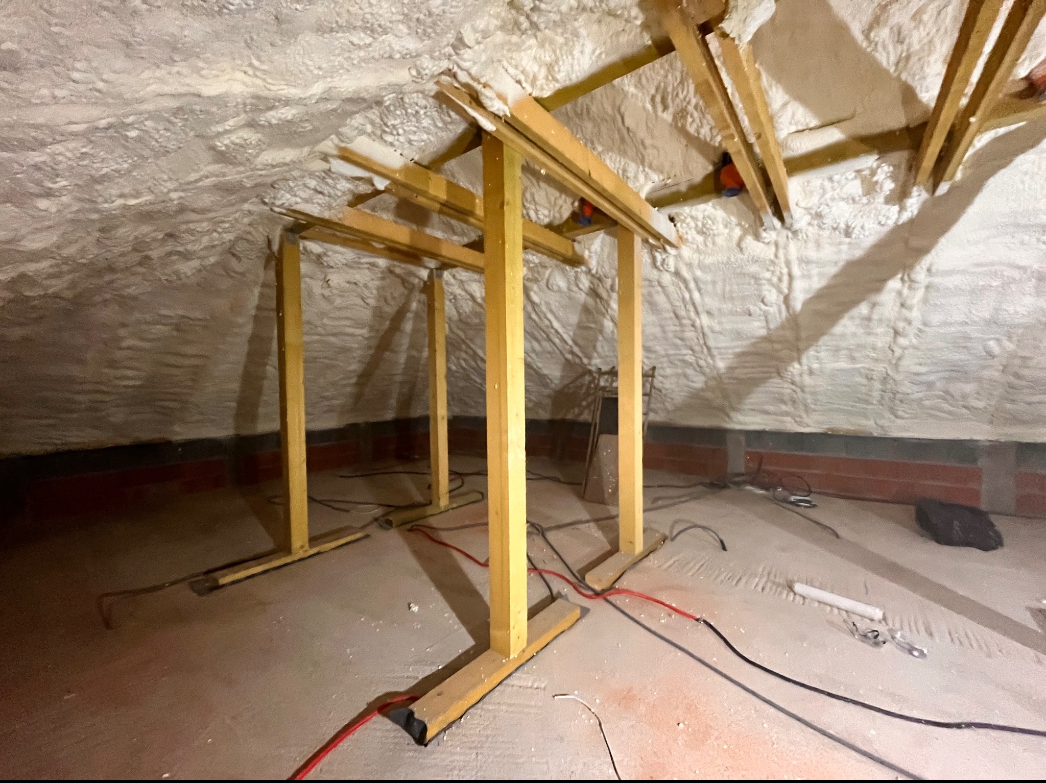 Poddasze z drewnianą konstrukcją i nałożoną białą pianką izolacyjną, widoczne przewody elektryczne na podłodze.