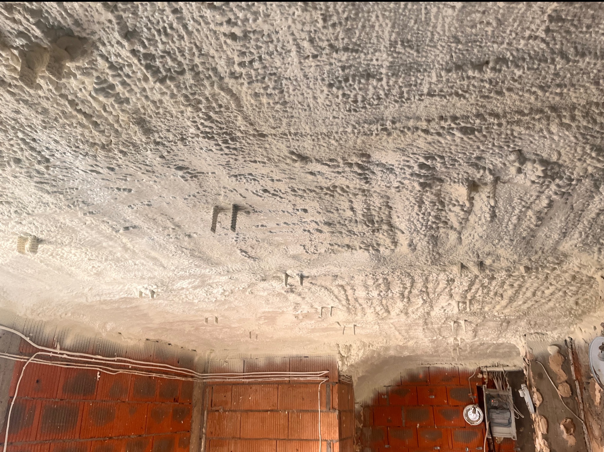 Świeżo nałożona, nierówna warstwa izolacji natryskowej na suficie i górnej części ścian budynku w trakcie budowy, widoczne przewody elektryczne i surowa cegła.