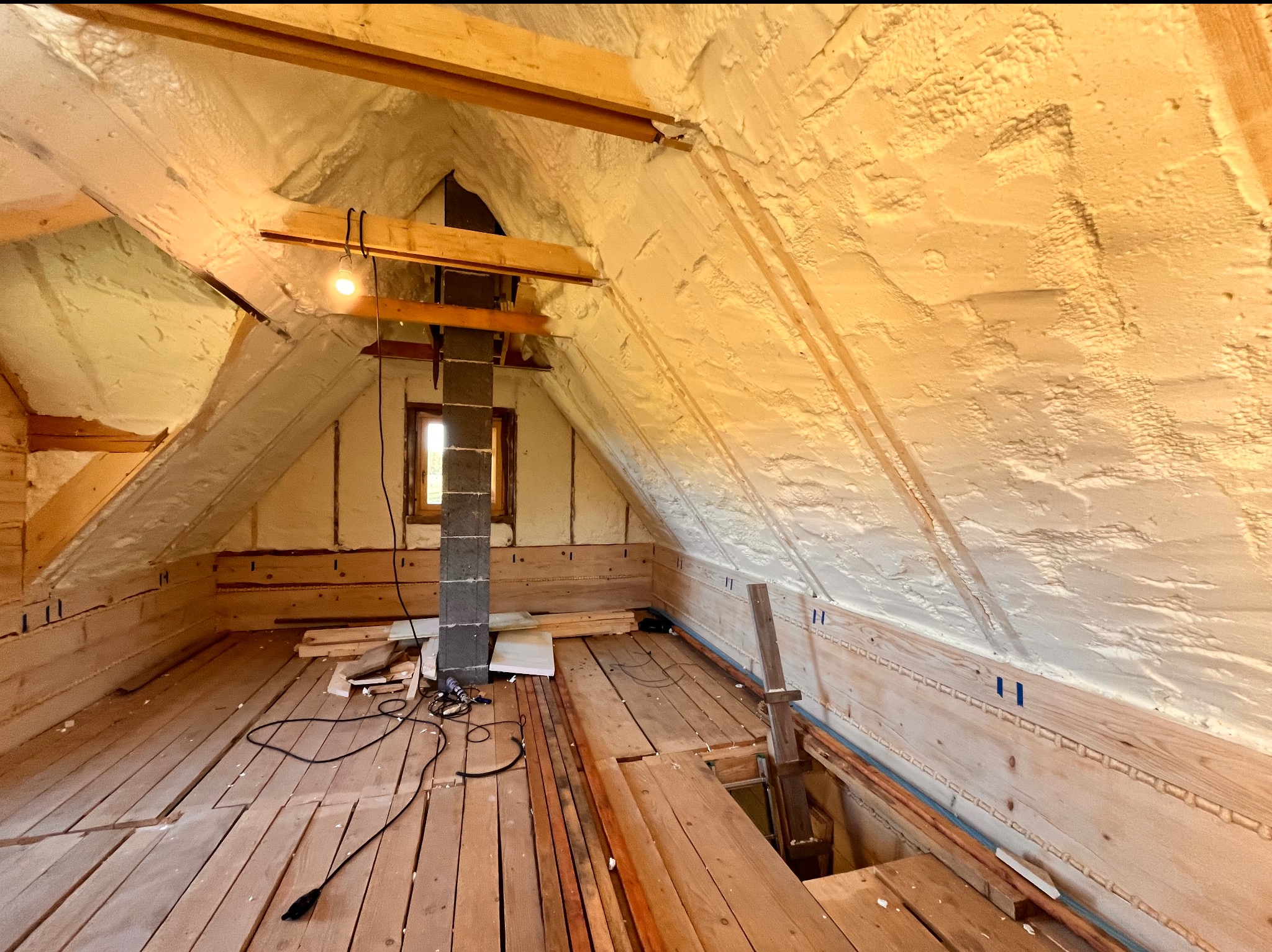 Poddasze w trakcie prac izolacyjnych, ściany i dach pokryte pianą, widoczne drewniane elementy konstrukcyjne, komin z cegły, kable, narzędzia i fragmenty drewnianej podłogi.