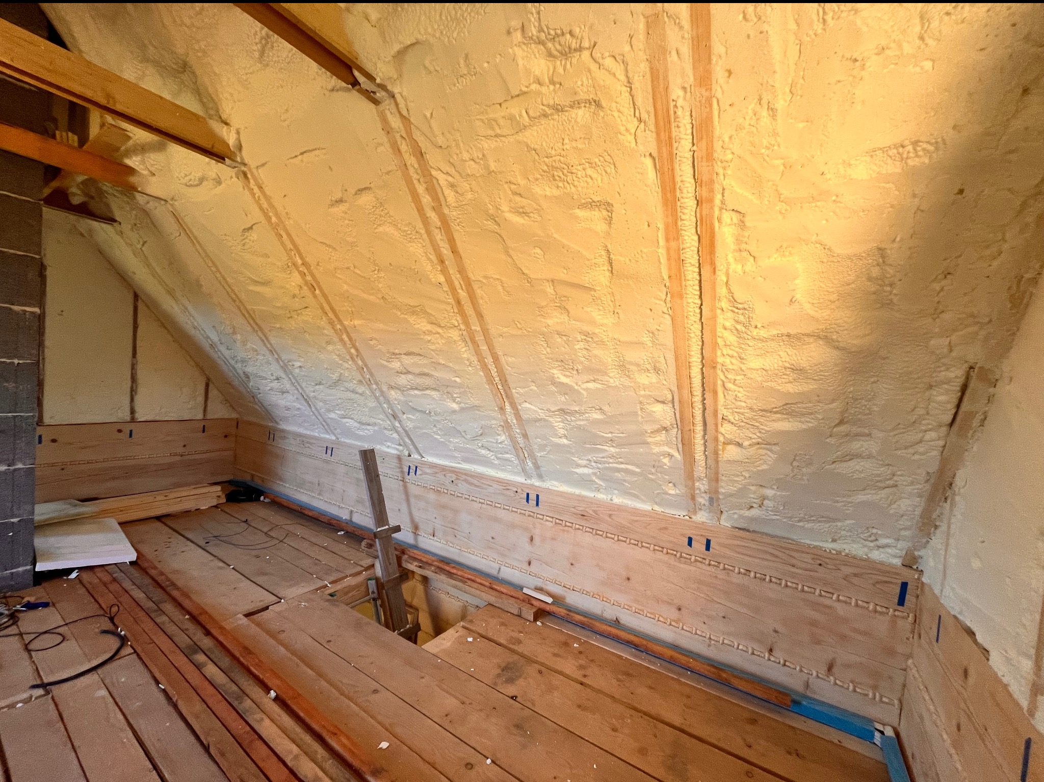 Poddasze w trakcie ocieplania pianką: widoczne drewniane elementy konstrukcyjne, fragment komina z szarego pustaka oraz świeżo nałożona warstwa izolacji z pianki o nierównej fakturze, częściowo...