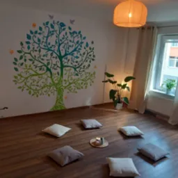Jasne wnętrze sali do medytacji z poduszkami na podłodze, ścianą ozdobioną malowidłem drzewa życia, roślinami w doniczkach i lampionem