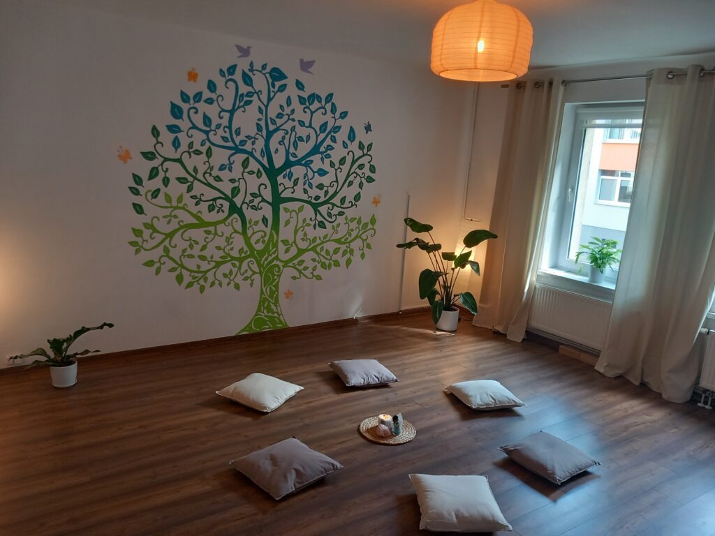 Jasne wnętrze sali do medytacji z poduszkami na podłodze, ścianą ozdobioną malowidłem drzewa życia, roślinami w doniczkach i lampionem