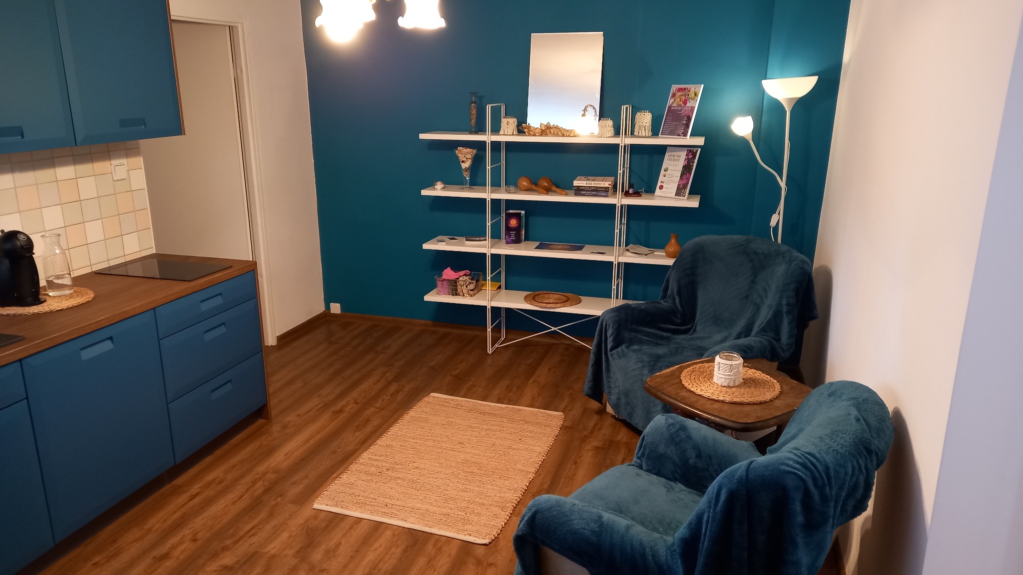 Gabinet terapii naturalnej: Dwa turkusowe fotele z pledami, stolik z dekoracją, biały regał z akcesoriami i materiałami informacyjnymi na tle turkusowej ściany, dywanik z juty na podłodze.