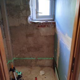 SEBUD Sebastian Niedużak - Remontowane pomieszczenie z oknem, gdzie zielony laser wyznacza poziomy na surowych ścianach i częściowo zamontowanych płytach gipsowo-kartonowych, widoczne narzędzia budowlane.