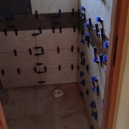 SEBUD Sebastian Niedużak - Układanie jasnych płytek ściennych z efektem marmuru w małym pomieszczeniu, prawdopodobnie łazience, z widocznymi klipsami poziomującymi i nieukończoną podłogą.