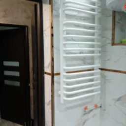 Biały, drabinkowy grzejnik łazienkowy na ścianie z marmurowymi płytkami, widoczny fragment bojlera Elprom i drzwi z czarną ościeżnicą.