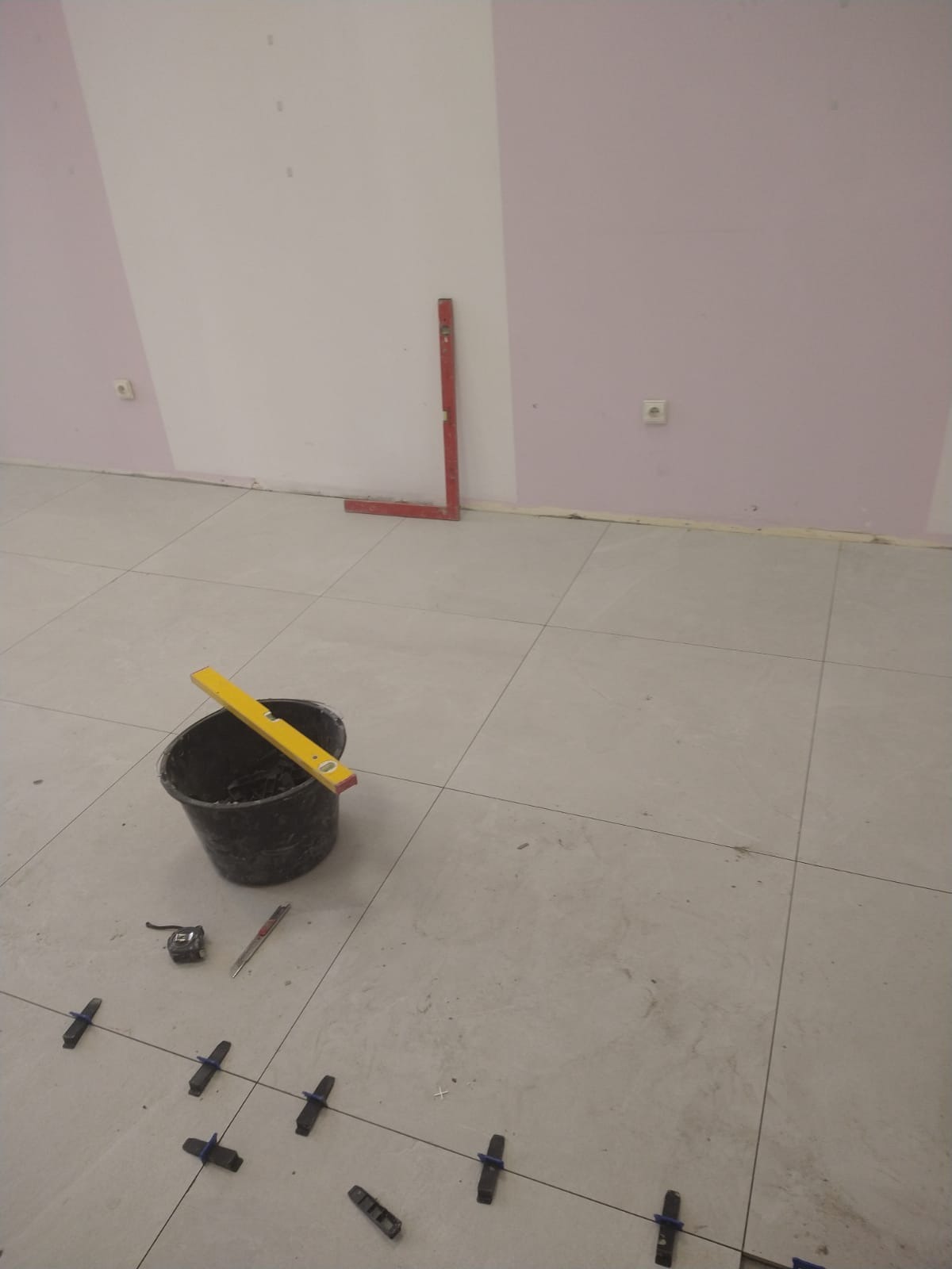 Układanie jasnych płytek podłogowych z użyciem krzyżyków dystansowych, widoczny kubeł z narzędziami i poziomica, ściana w dwóch odcieniach różu i bieli.