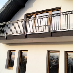 Łukasz Wolszczak - Balkon z metalową balustradą o wzorze greckim na tle białej elewacji domu z drewnianymi oknami. Ujęcie z dołu, widoczne odbicia drzew w szybach.