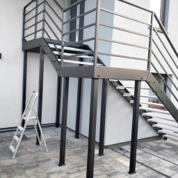 Łukasz Wolszczak - Zewnętrzne schody metalowe z ażurowymi stopniami i prostą balustradą w kolorze antracytowym, wsparte na słupach. W tle biała ściana budynku i drabina.