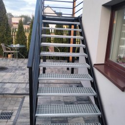 Łukasz Wolszczak - Zewnętrzne schody metalowe z kratowymi stopniami prowadzą w górę, przylegając do ściany budynku. Widoczne elementy balustrady i otoczenie z kostki brukowej.