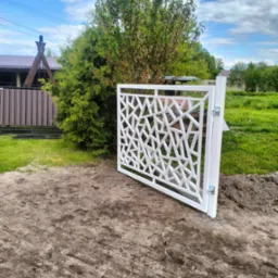Biała, metalowa brama wjazdowa z geometrycznym wzorem, zamontowana na tle zieleni. Widoczne zawiasy i fragment domu w tle.