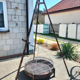 Metalowy trójnóg z rusztem do grillowania na zewnątrz, ustawiony na kostce brukowej przed budynkiem z białej cegły i stertami materiałów budowlanych w tle.