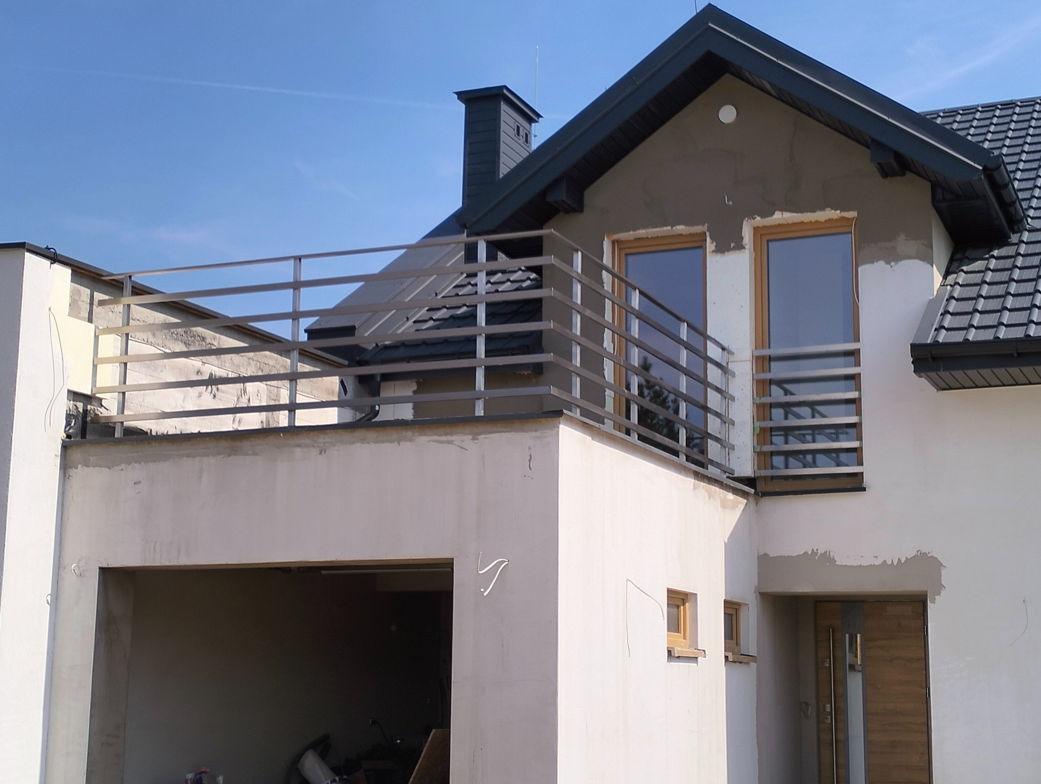 Nowoczesna balustrada balkonowa z poziomych profili na tle surowej elewacji domu w trakcie budowy. Proste, metalowe barierki przy oknach.