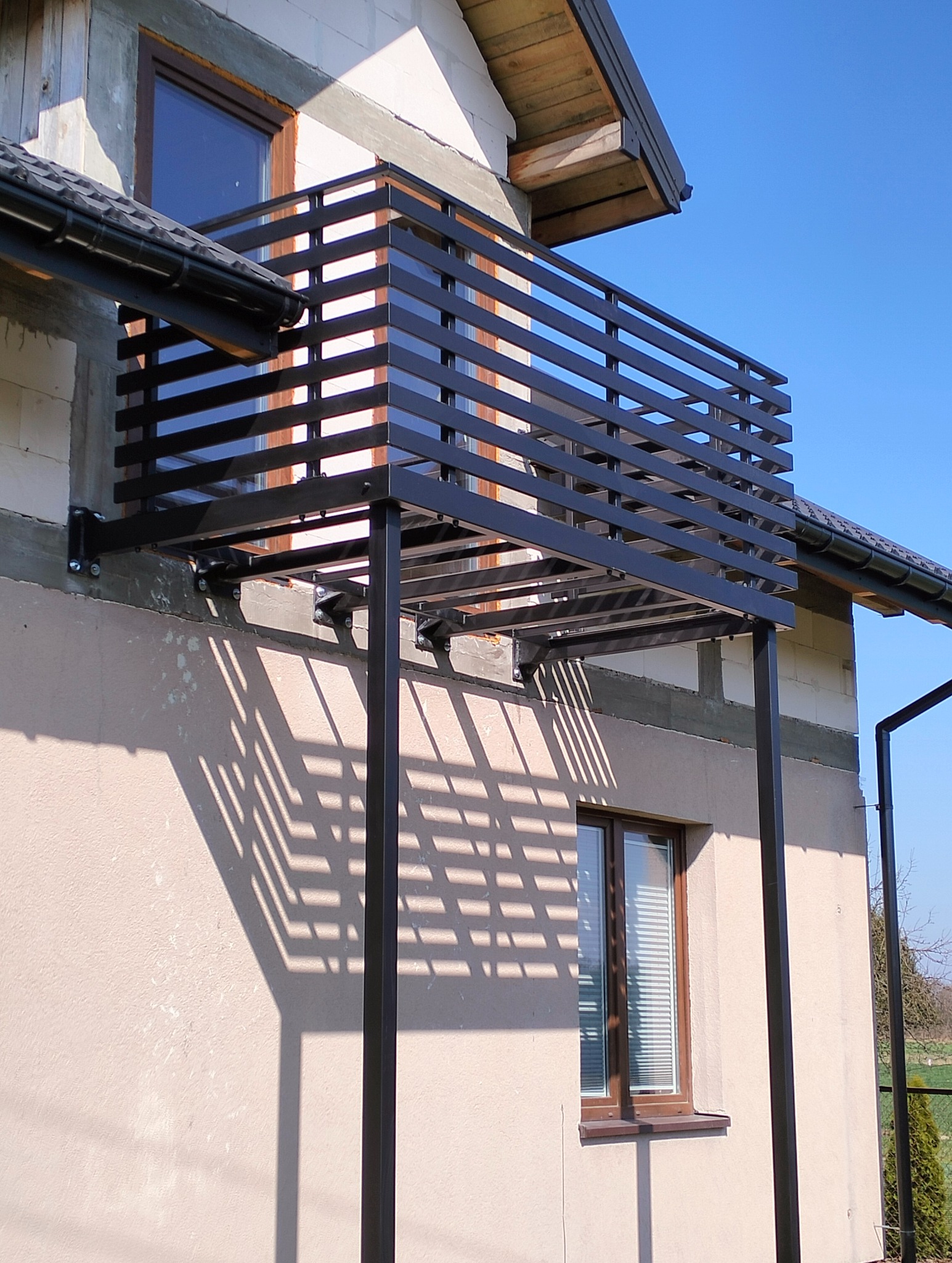 Nowoczesna, czarna balustrada balkonowa z poziomych profili, zamontowana do elewacji budynku. Widoczne detale mocowania i cień balustrady na ścianie.