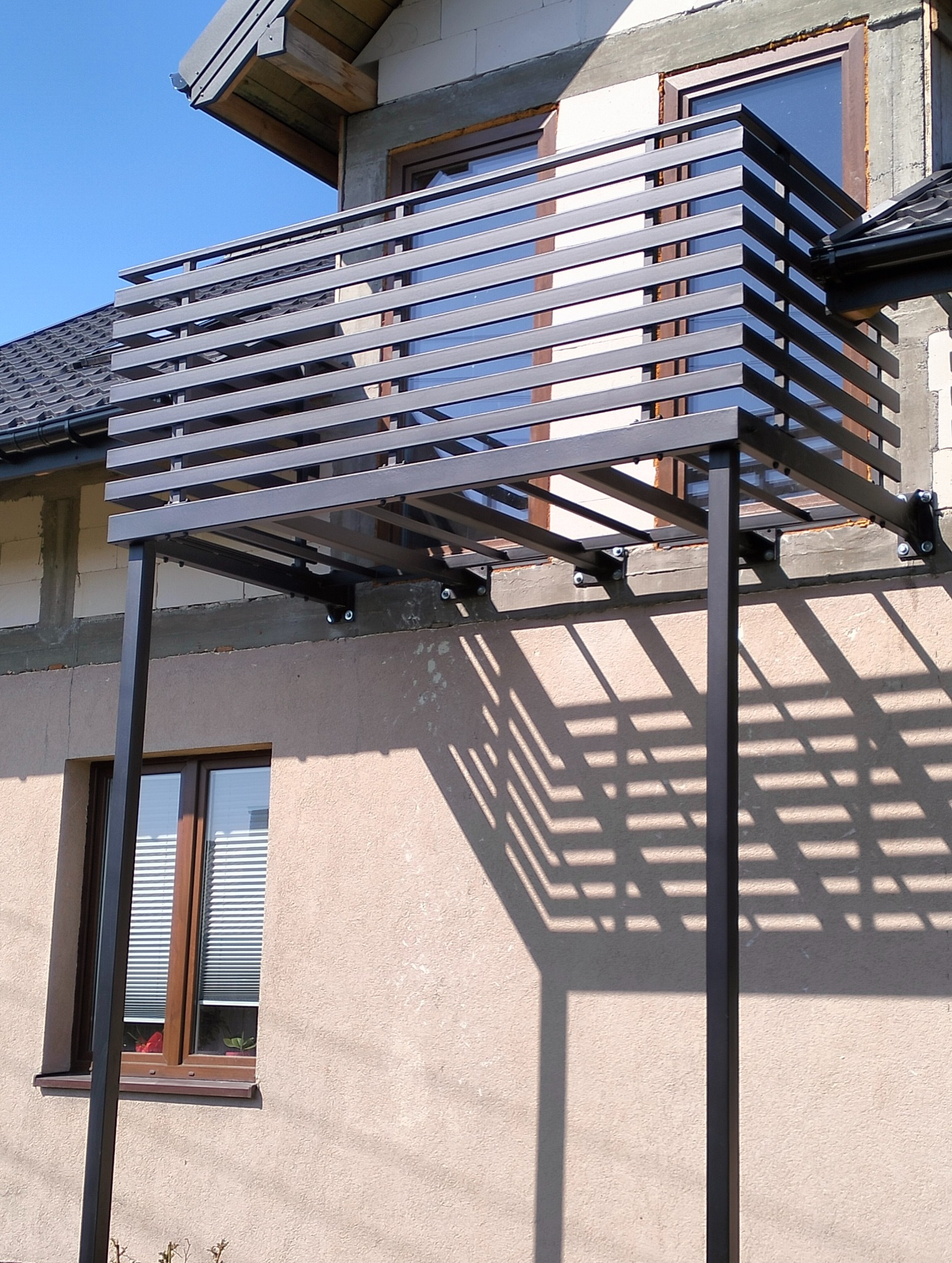 Nowoczesna, metalowa balustrada balkonowa z poziomymi szczeblami, zamontowana na elewacji budynku. Widoczne mocowania i cień konstrukcji.