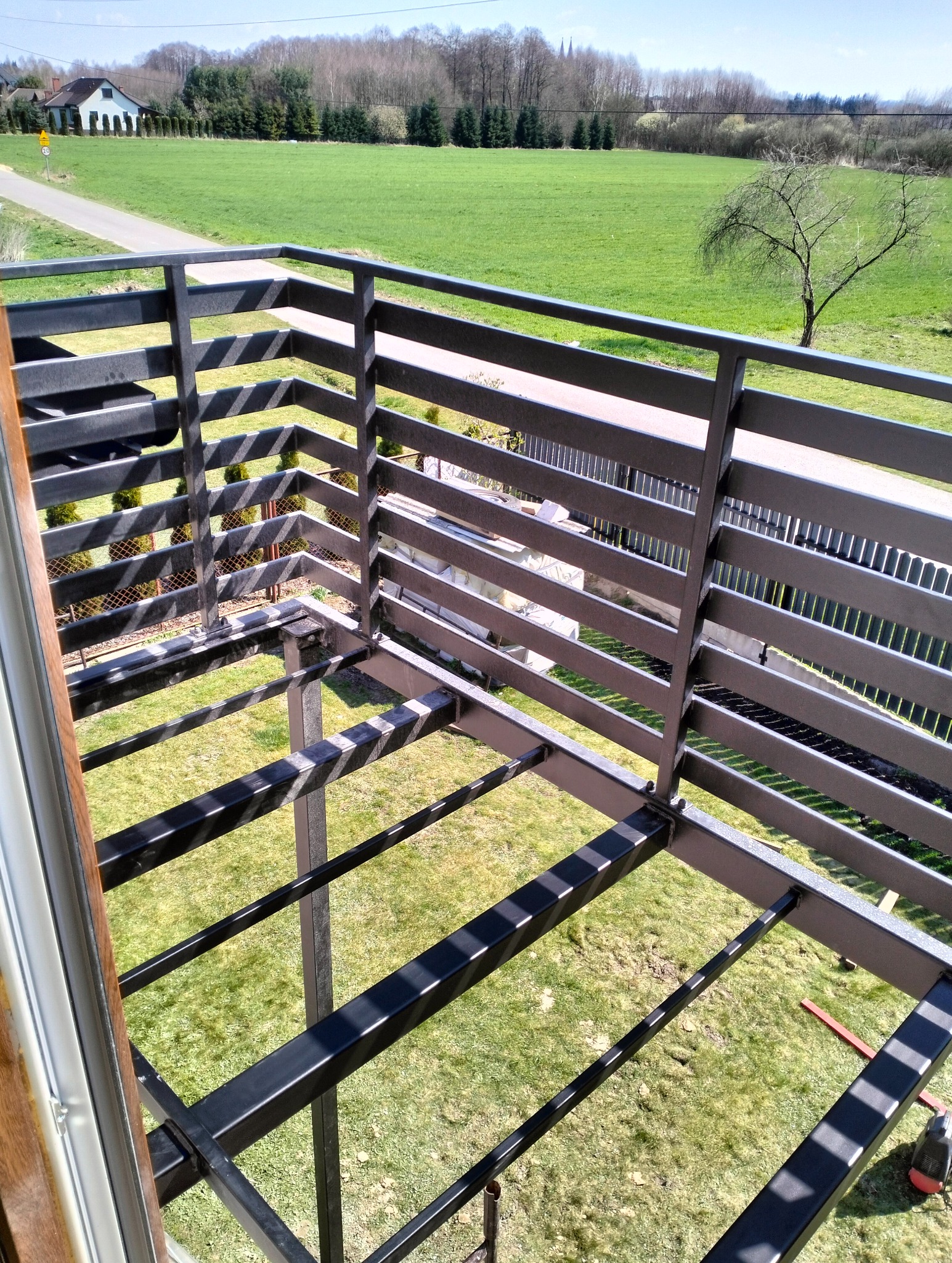 Surowa, metalowa konstrukcja balustrady balkonowej z widokiem na zielony krajobraz. Widoczne spawy i elementy mocujące, sugerujące etap montażu.