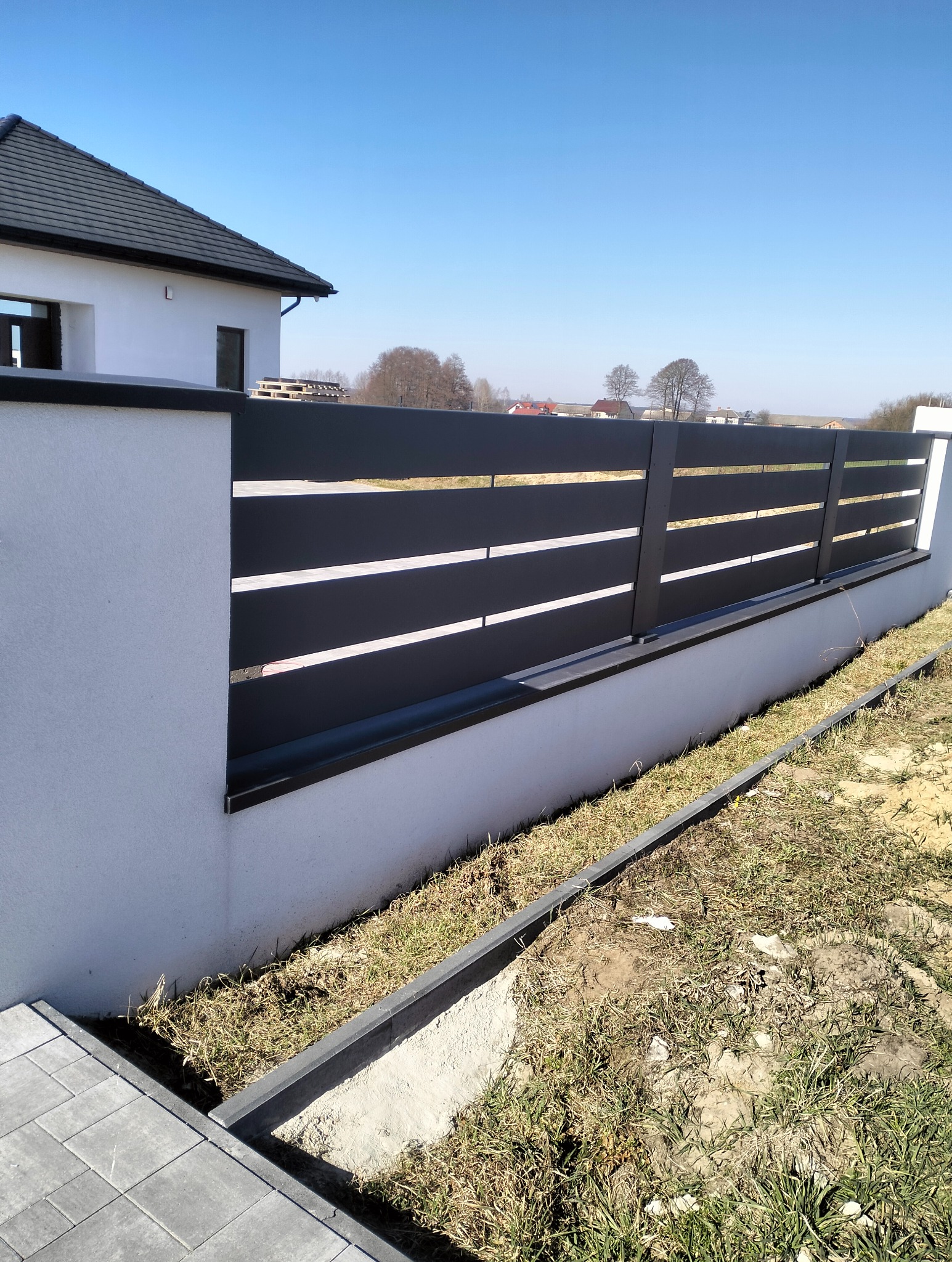 Nowoczesne ogrodzenie panelowe w kolorze grafitowym z poziomymi przerwami, zamontowane na niskim, murowanym murku. W tle dom jednorodzinny i błękitne niebo.