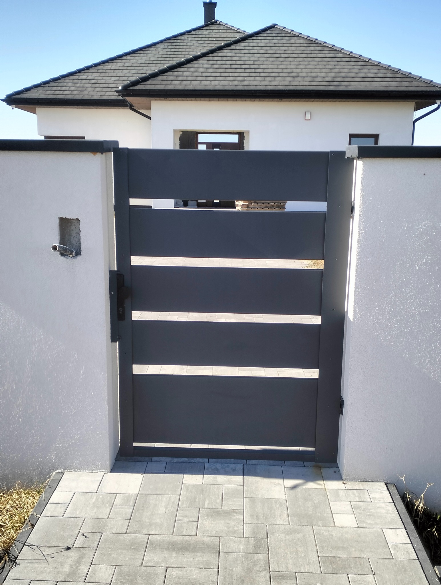 Nowoczesna, pozioma brama wjazdowa w kolorze grafitowym, zamontowana pomiędzy białymi słupkami. W tle widoczny dom z szarym dachem i jasną elewacją.