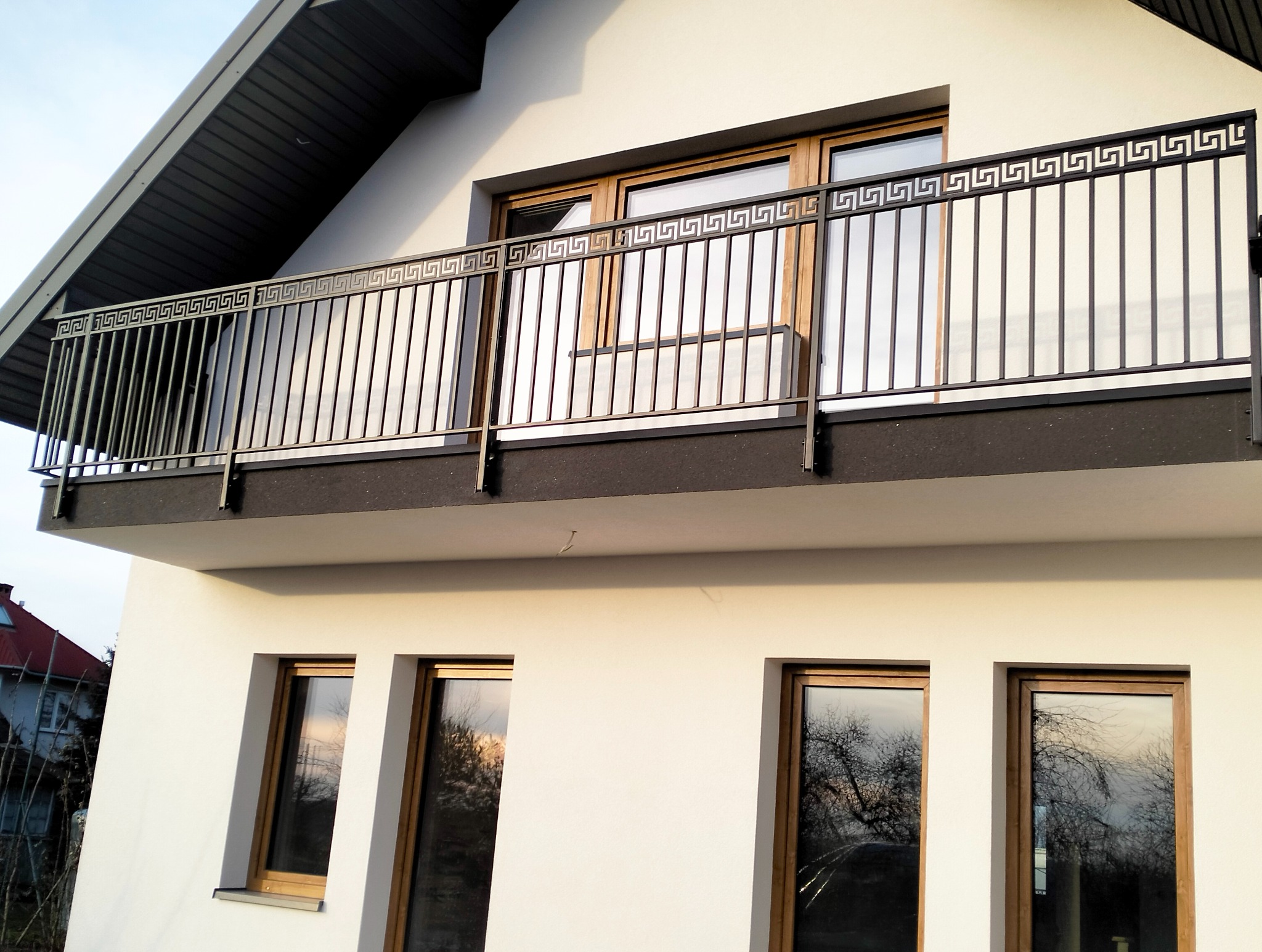 Balkon z metalową balustradą o wzorze greckim na tle białej elewacji domu z drewnianymi oknami. Ujęcie z dołu, widoczne odbicia drzew w szybach.