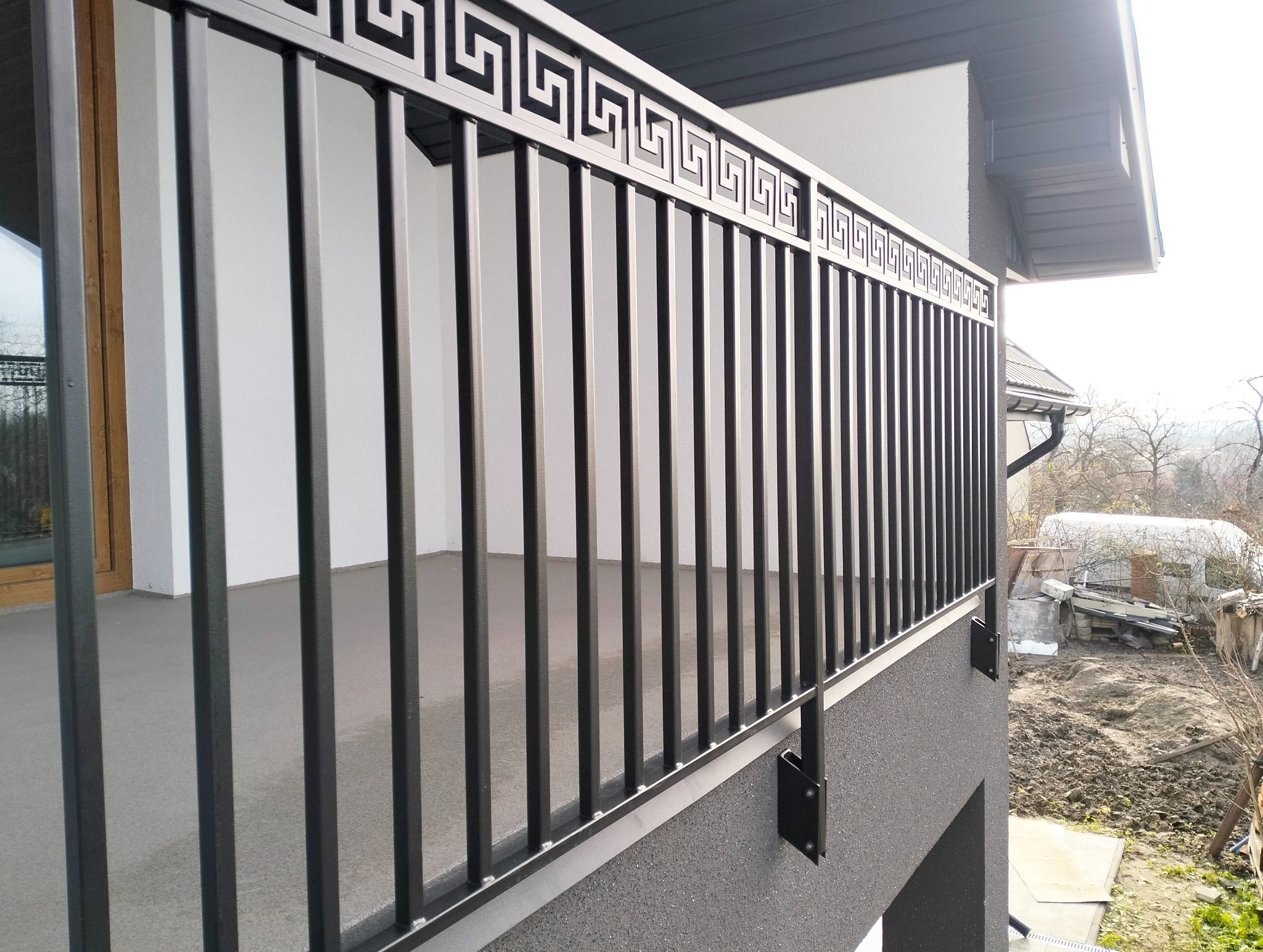Czarna balustrada balkonowa z motywem greckim, zamontowana na szarym balkonie, z widokiem na otoczenie.