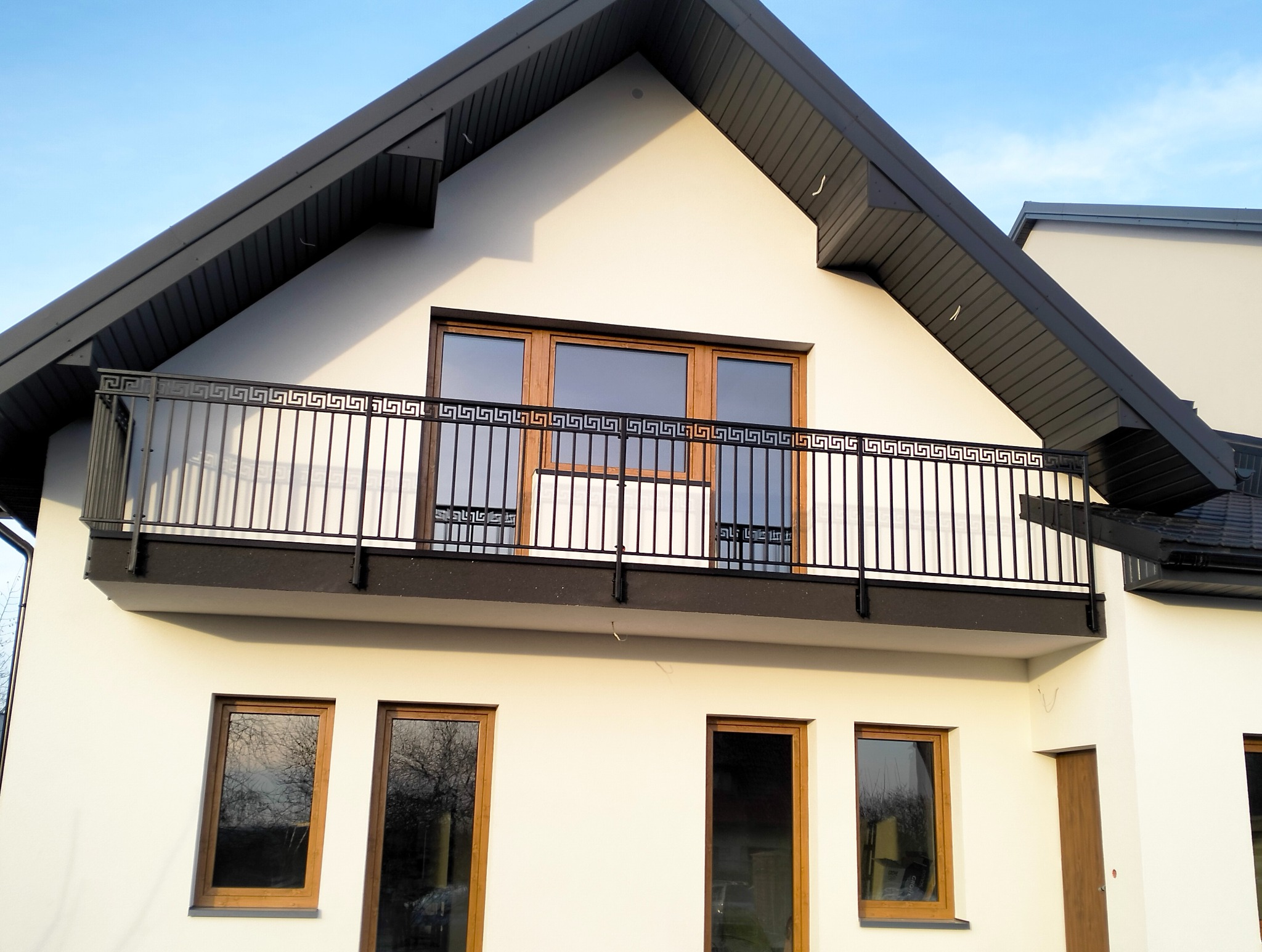 Czarna, metalowa balustrada balkonowa z motywem greckim na tle białej elewacji domu i błękitnego nieba. Widok z dołu, podkreślający detale wzoru.
