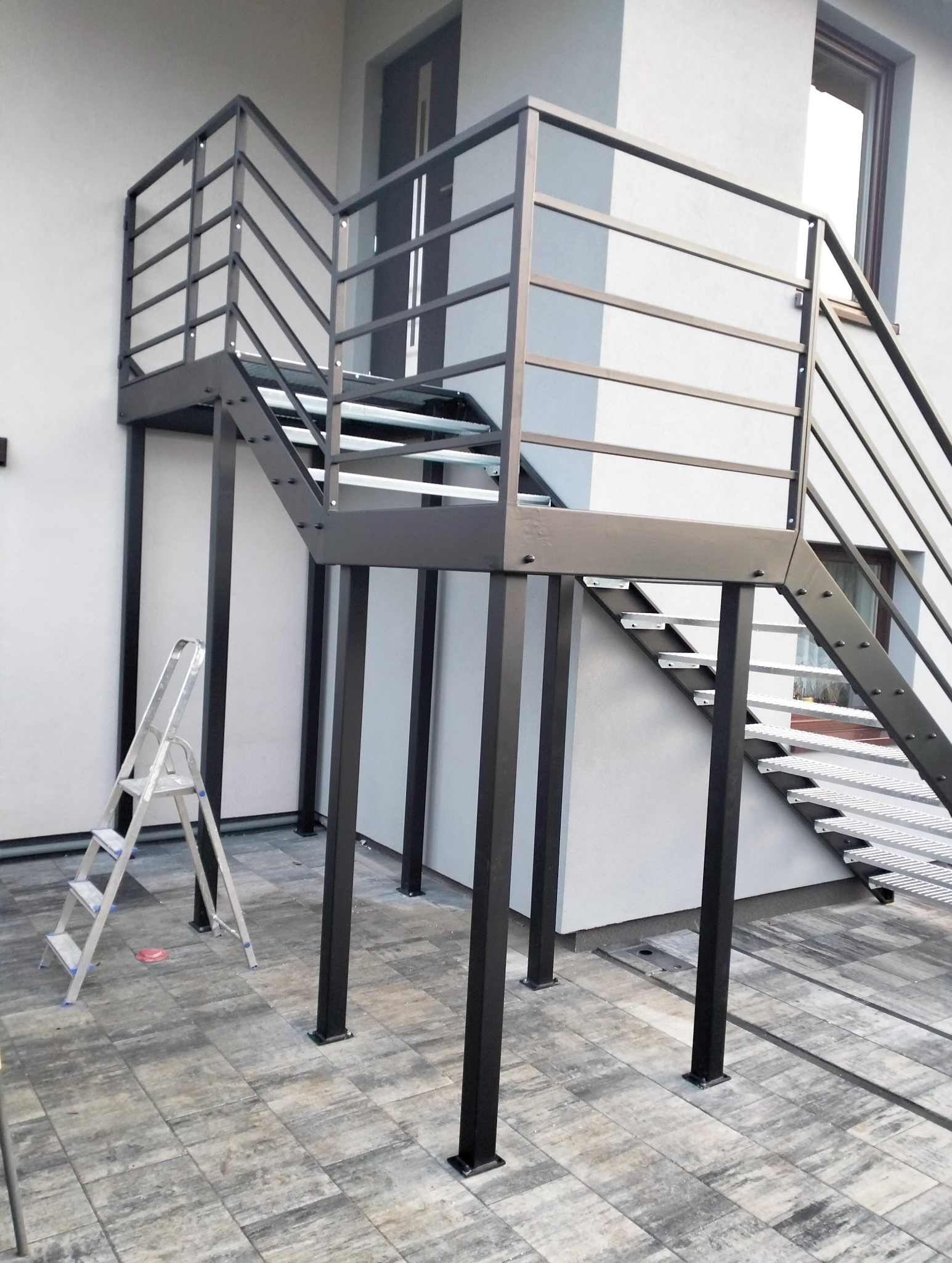 Zewnętrzne schody metalowe z ażurowymi stopniami i prostą balustradą w kolorze antracytowym, wsparte na słupach. W tle biała ściana budynku i drabina.