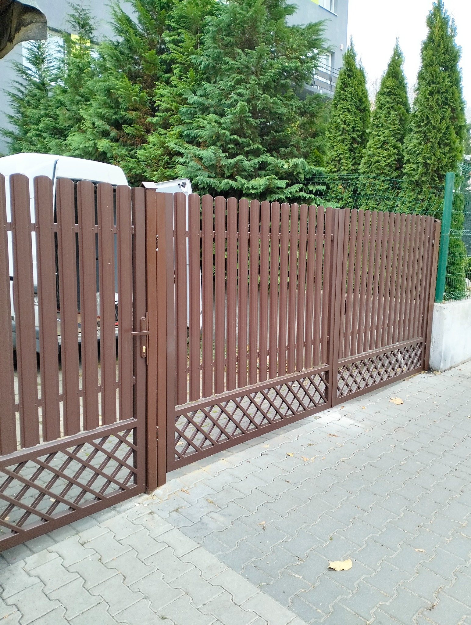 Brązowa brama wjazdowa z pionowych sztachet i dekoracyjnym, ażurowym dołem, na tle zielonych drzew i fragmentu białego vana, widziana z poziomu chodnika.