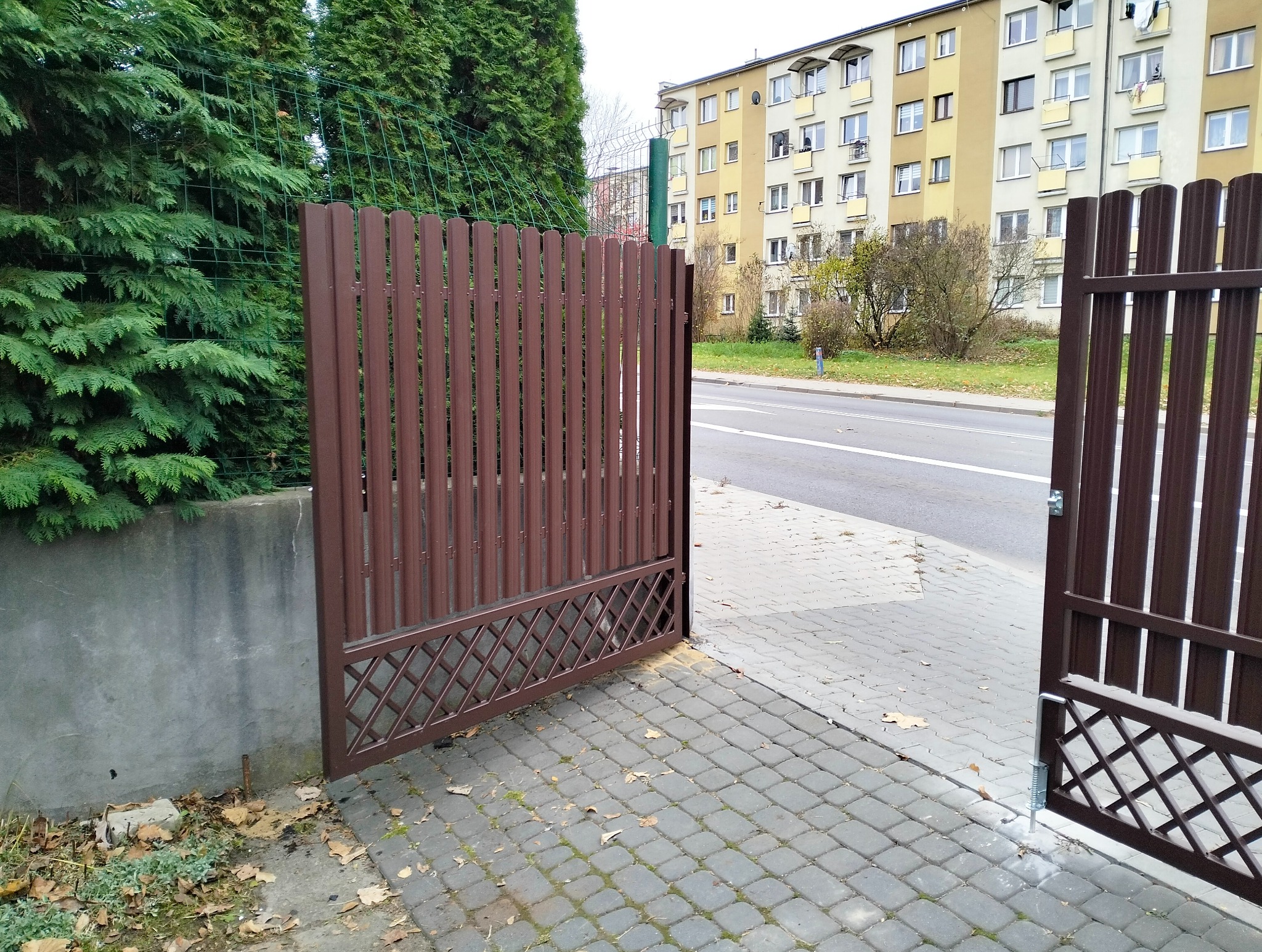 Otwarta, brązowa brama wjazdowa z metalowymi sztachetami i dekoracyjnym wzorem kratki w Tczowie, na tle chodnika z kostki i zieleni. Widok z zewnątrz posesji.