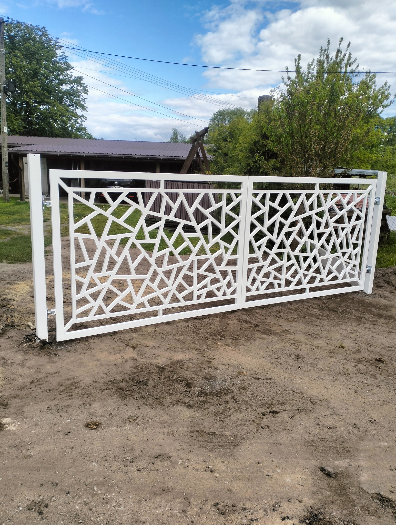 Biała, metalowa brama wjazdowa z geometrycznym wzorem na tle zieleni i błękitnego nieba. Nowoczesny design bramy.