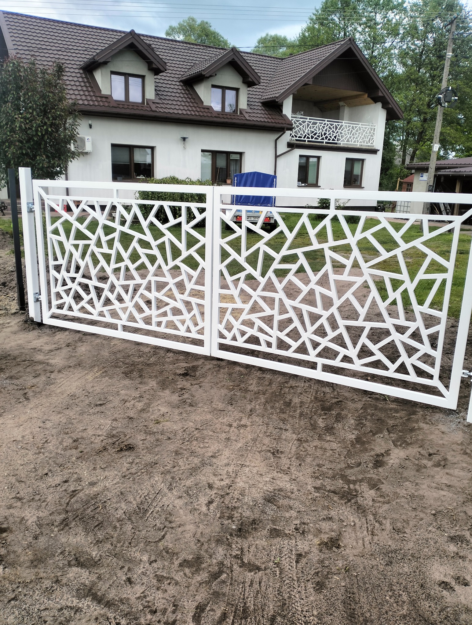 Nowoczesna, biała brama wjazdowa z geometrycznym wzorem na tle domu z balkonem, widziana z perspektywy ziemi.