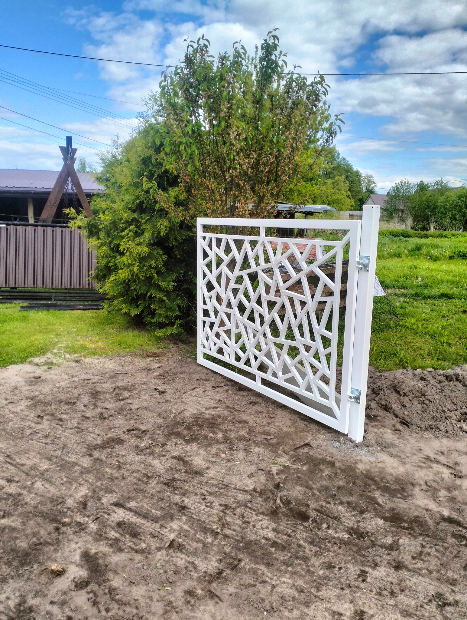 Biała, metalowa brama wjazdowa z geometrycznym wzorem, zamontowana na tle zieleni. Widoczne zawiasy i fragment domu w tle.