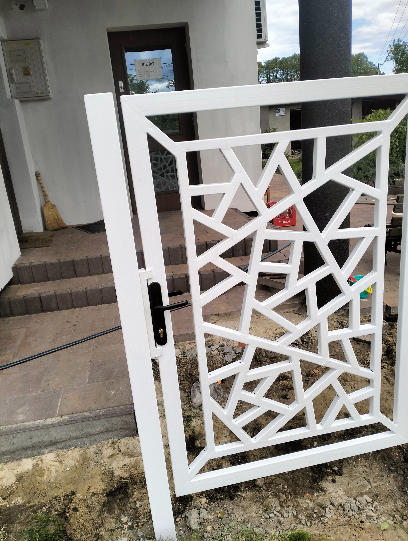 Biała, nowoczesna furtka z geometrycznym wzorem, czarna klamka, widok na schody i budynek. Furtka w trakcie montażu, ziemia wokół.
