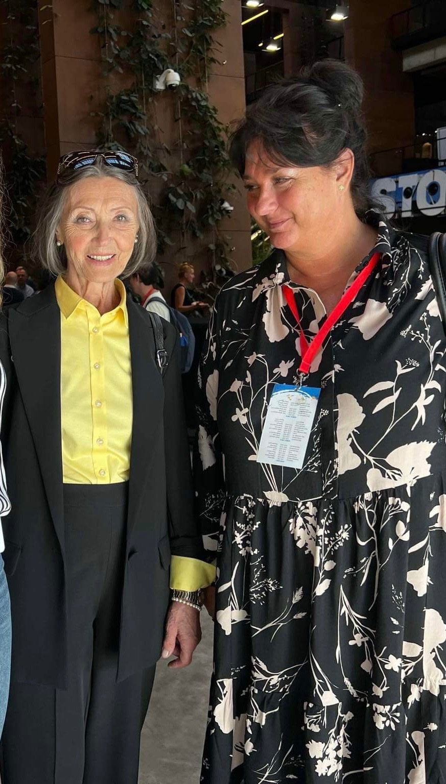 Dwie kobiety podczas konferencji, jedna w czarnym garniturze z żółtą koszulą, druga w kwiecistej sukience z identyfikatorem na szyi. Uśmiechnięte, w tle rośliny.