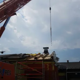 Czerwony dźwig podnosi element dachu z częściowo rozebranego budynku, widoczne rusztowania i gruz.