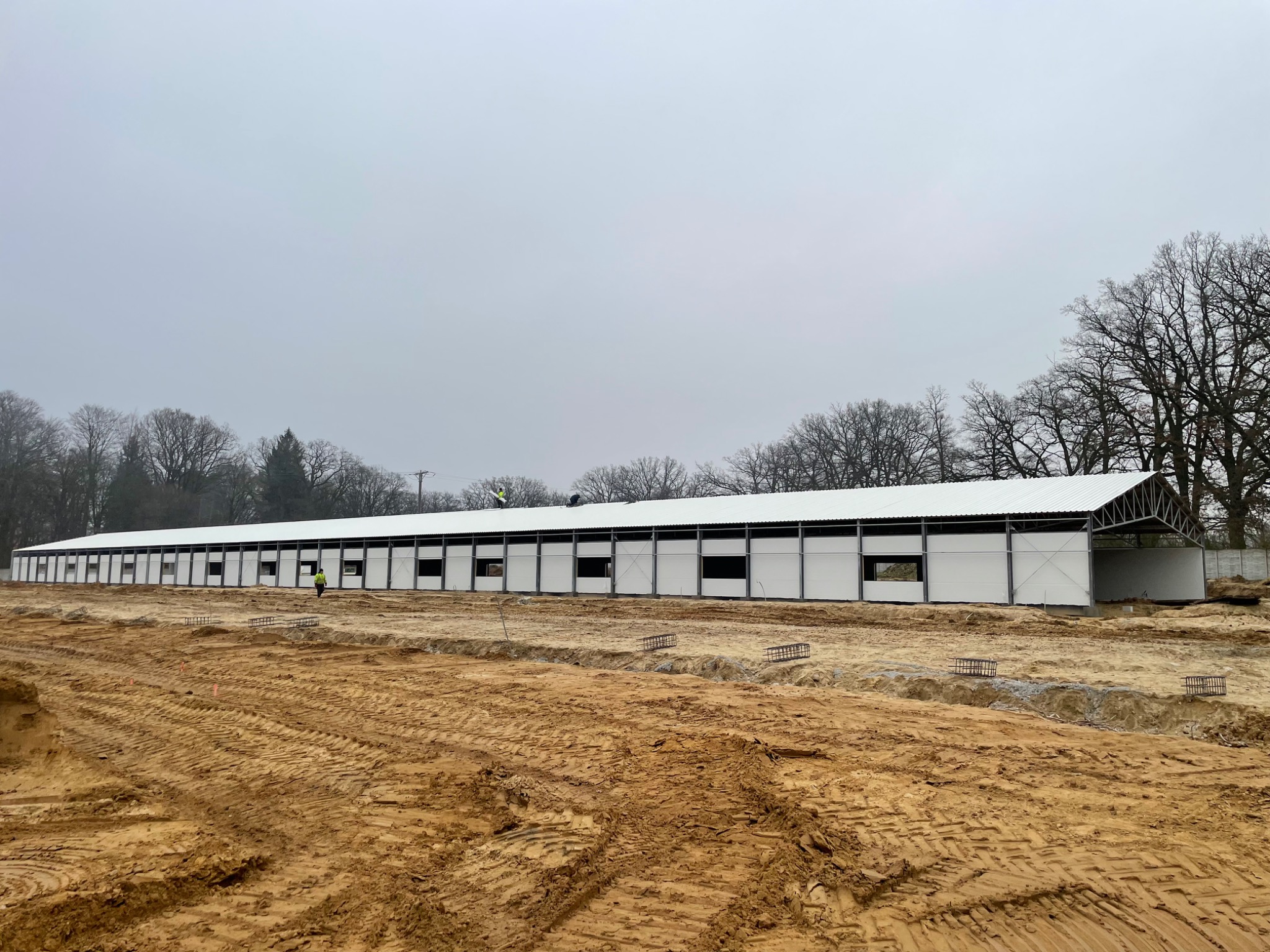 Budowa hali z płyt warstwowych w trakcie realizacji, widoczne przygotowane fundamenty i pracownik w odblaskowej kamizelce na tle konstrukcji i pochmurnego nieba.