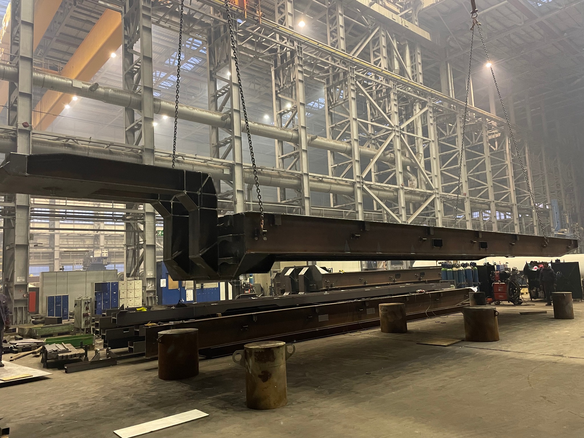 Surowa stalowa konstrukcja hali podnoszona łańcuchami suwnicy w hali produkcyjnej, widoczne spawarki i butle z gazem.