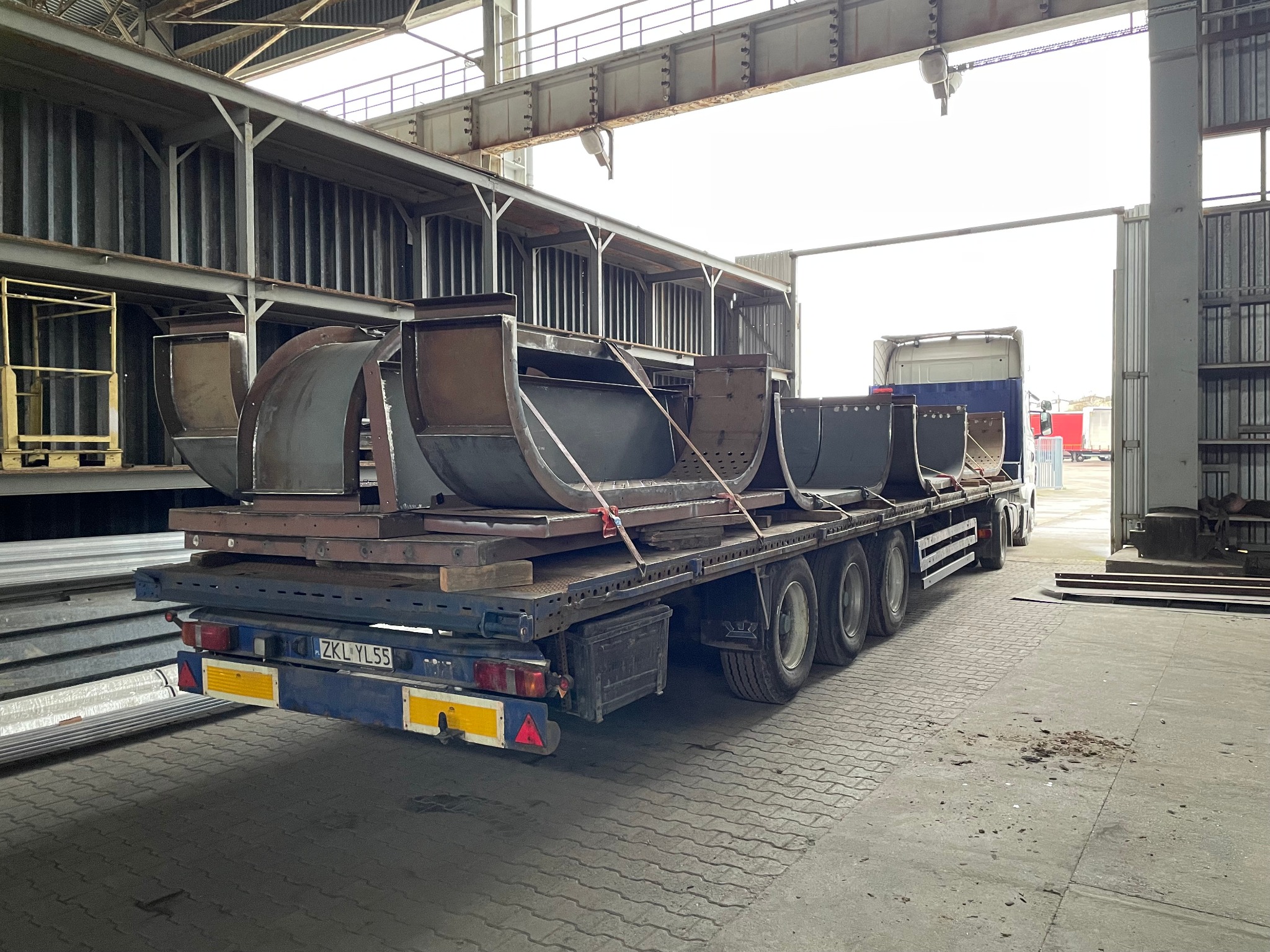 Transport elementów konstrukcji stalowej, prawdopodobnie fragmentów rurociągu, na naczepie ciężarówki. Widoczne spawane łączenia i mocowania. Hala produkcyjna w tle.