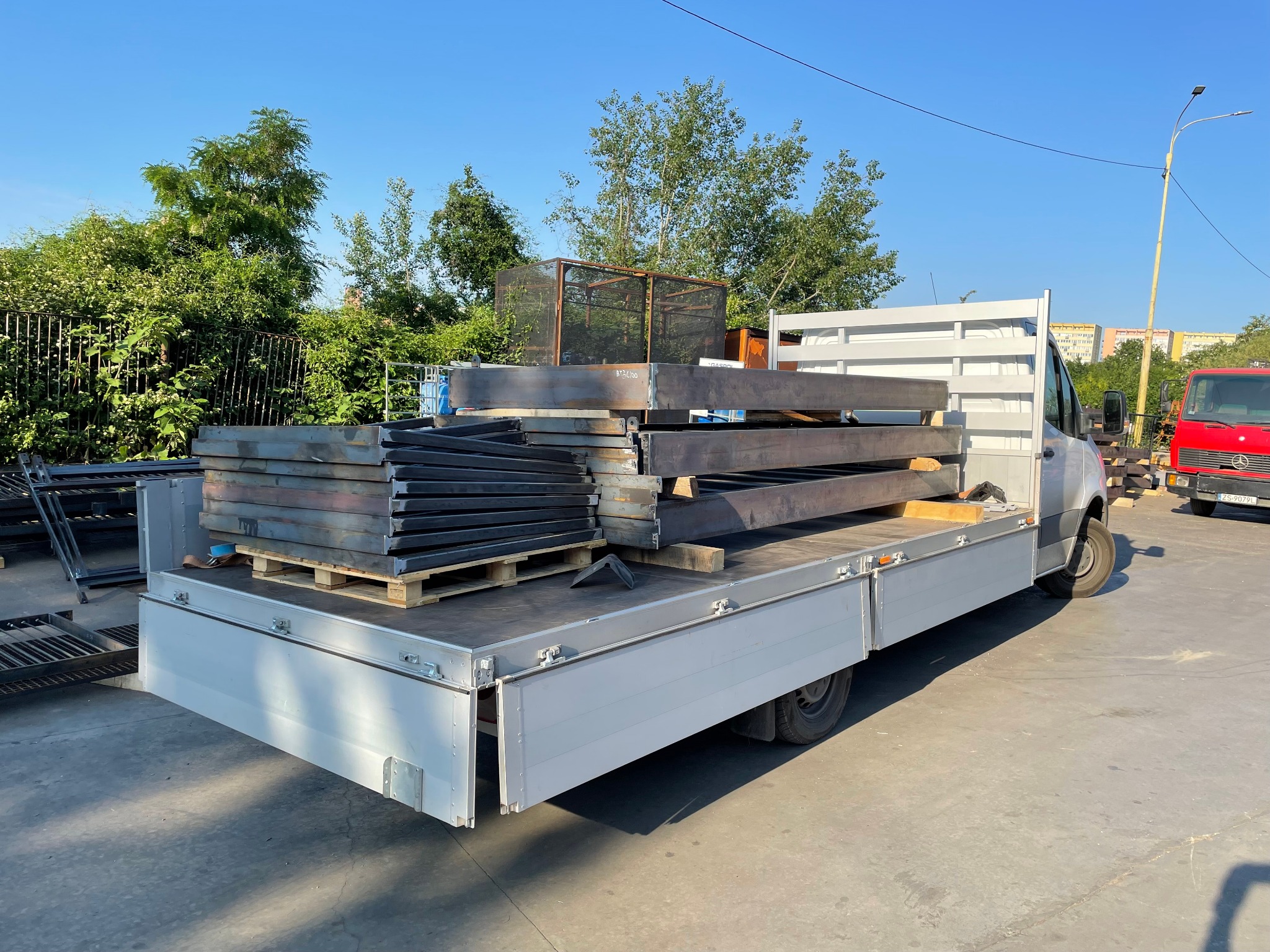 Ciężarówka z platformą załadowana stalowymi elementami konstrukcyjnymi, w tle zieleń i budynki.