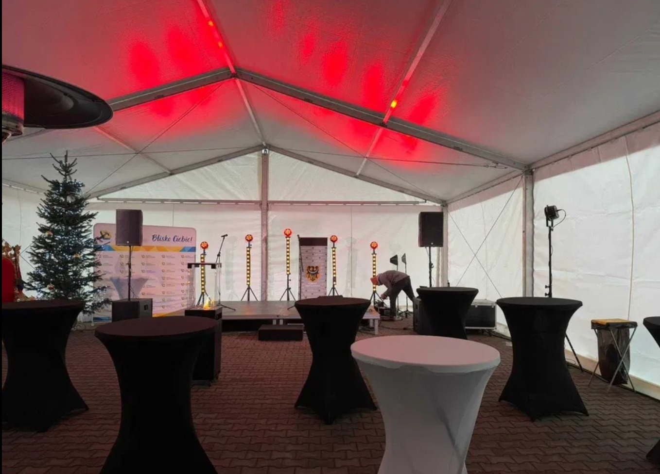 Wnętrze hali namiotowej z oświetleniem, stołami koktajlowymi, sceną i choinką. Osoba konfiguruje sprzęt.