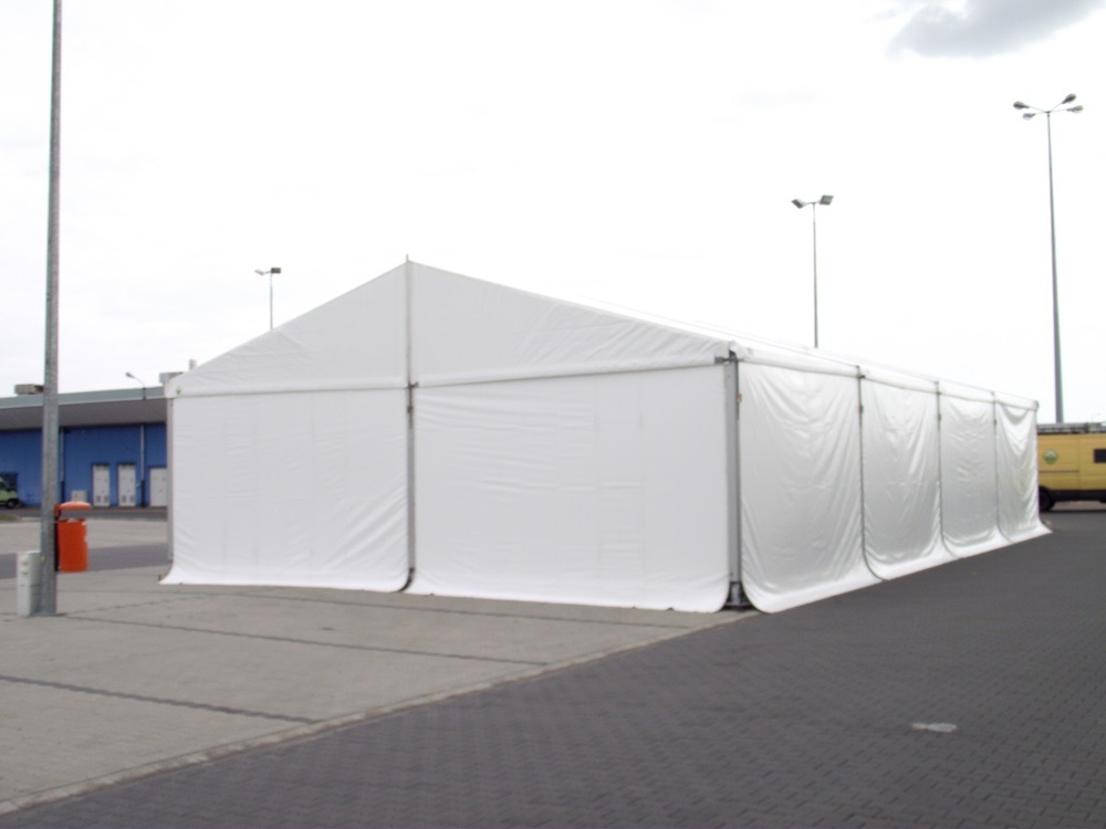 Biała hala namiotowa z widocznymi stalowymi elementami konstrukcyjnymi, ustawiona na betonowym placu przy budynku. Widoczne słupy oświetleniowe i fragment pojazdu.