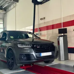 Szary samochód Audi Q7 na czerwonym podnośniku w jasnym warsztacie z widocznym sprzętem do ustawiania geometrii kół.