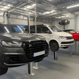 Wnętrze warsztatu samochodowego z trzema pojazdami Audi na podnośnikach: szary SUV, biały van i czerwony kabriolet.