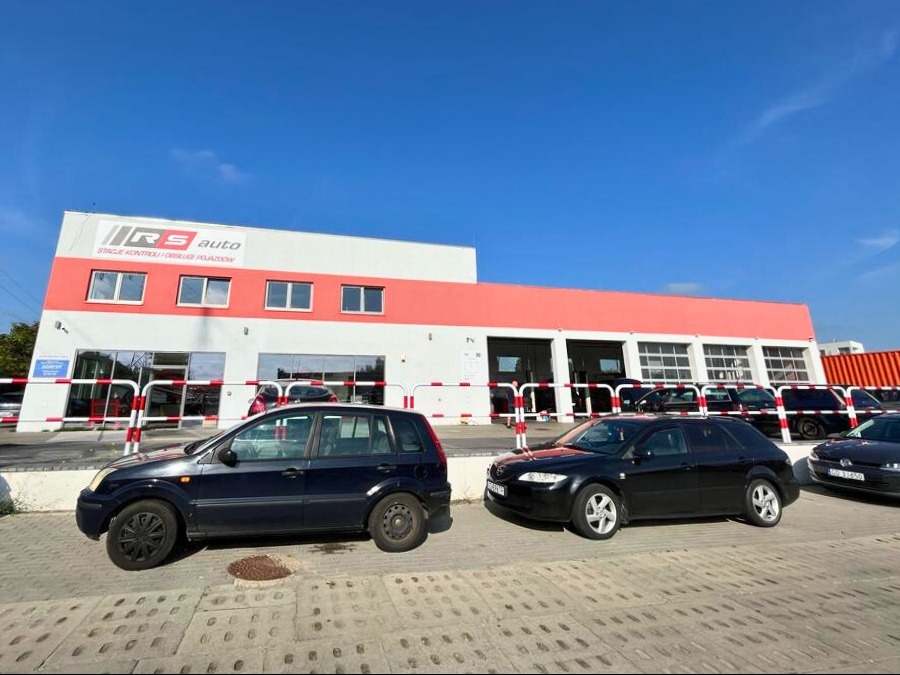 Budynek warsztatu samochodowego RS Auto w Gdańsku z zaparkowanymi samochodami przed wjazdem do hali. Widoczny szyld firmy z nazwą i hasłem 'Stacje kontroli i obsługi pojazdów'.