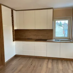 Nowoczesna kuchnia z białymi, lakierowanymi frontami i drewnopodobnym blatem oraz elementami wykończeniowymi, widok na ulicę przez okno.