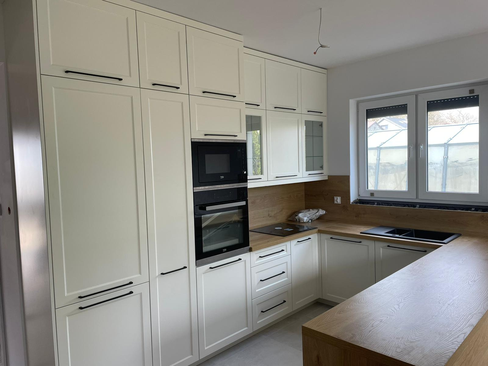 Jasna, nowoczesna kuchnia z kremowymi szafkami z frezowanymi frontami i długimi, czarnymi uchwytami, zabudowany piekarnik i mikrofalówka, blat imitujący drewno, okno z roletami.
