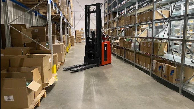 Wnętrze magazynu z regałami wypełnionymi kartonami i wózkiem widłowym. Perspektywa z dołu, oświetlenie sufitowe, betonowa podłoga.