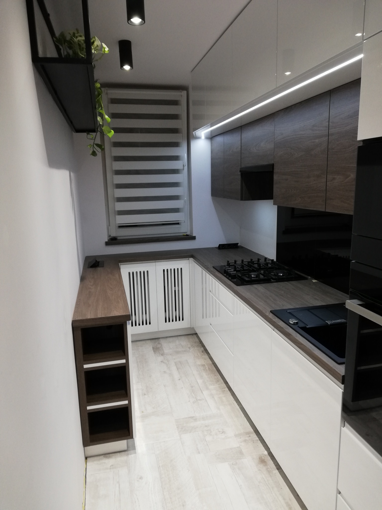 Nowoczesna kuchnia w kształcie litery U z białymi frontami dolnych szafek i górnymi szafkami w kolorze ciemnego drewna, oświetlenie LED pod szafkami, blaty w kolorze ciemnego drewna, płyta gazowa...