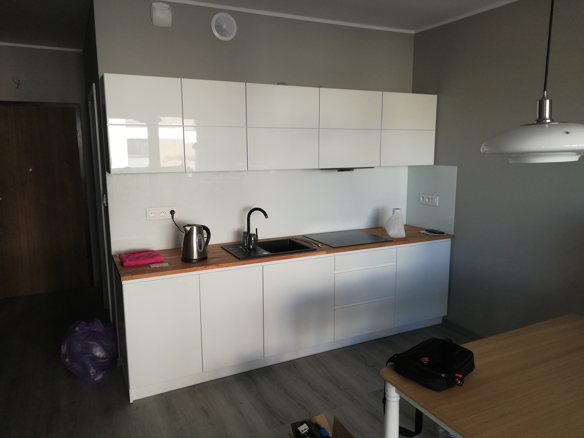 Nowoczesna kuchnia z białymi szafkami i drewnianym blatem, czarny zlew i płyta indukcyjna, minimalistyczne oświetlenie, widoczny czajnik elektryczny i torba na stole.