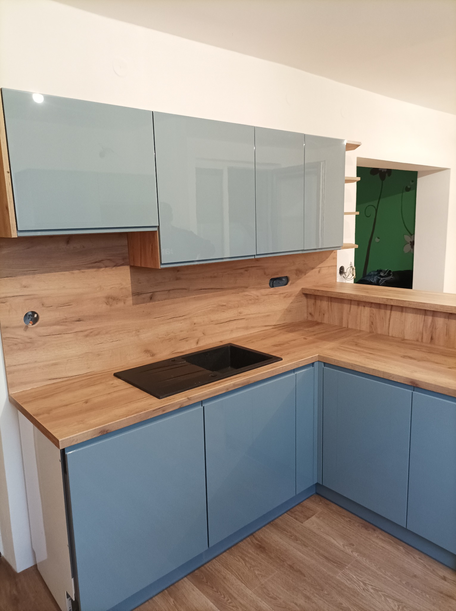 Niebieska kuchnia na wymiar z drewnianym blatem i czarnym zlewem, widoczne gniazdka elektryczne w ścianie, w tle zielona ściana z motywem roślinnym.