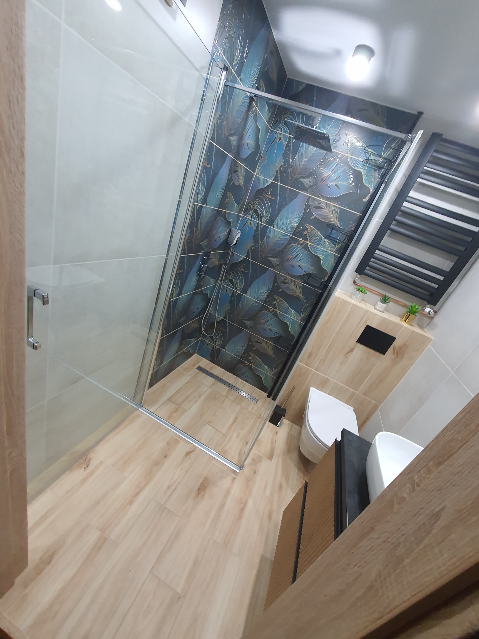 Nowoczesna łazienka z kabiną prysznicową ze szklanymi drzwiami, płytkami w odcieniach granatu i złota z motywem liści, drewnopodobną podłogą i grzejnikiem drabinkowym w kolorze czarnym.